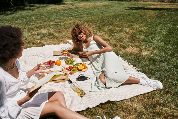 mujer afroamericana con teléfono inteligente y copa de vino hablando con su novia en el picnic de verano - Foto, Imagen