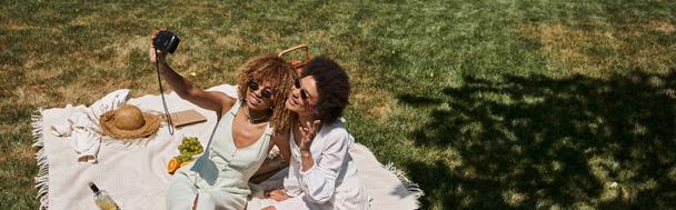 Neşeli Afrikalı Amerikalı kız arkadaşlar parktaki meyvelerin yanındaki battaniyenin üzerinde selfie çekiyorlar. - Fotoğraf, Görsel