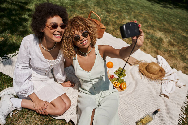 novias afroamericanas despreocupadas en gafas de sol tomando selfie en la cámara vintage, picnic, pancarta - Foto, Imagen