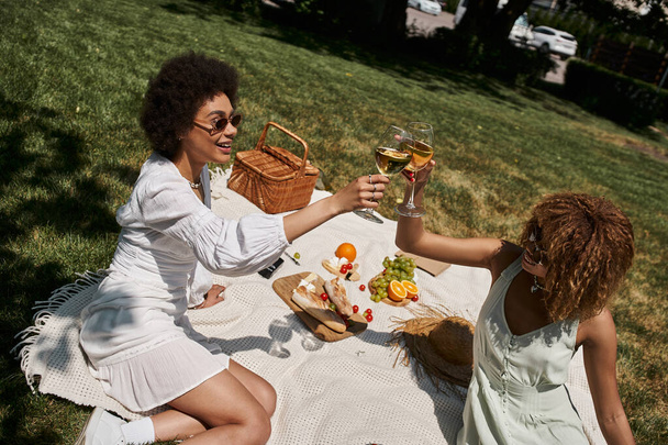 happy african american girlfriends clinking wine glasses on blanket and lawn in summer park - Fotografie, Obrázek