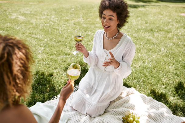 συναισθηματική Αφροαμερικανή γυναίκα με ποτήρι κρασιού μιλώντας με τη φίλη κατά τη διάρκεια του καλοκαιρινού πικ-νικ στο πάρκο - Φωτογραφία, εικόνα
