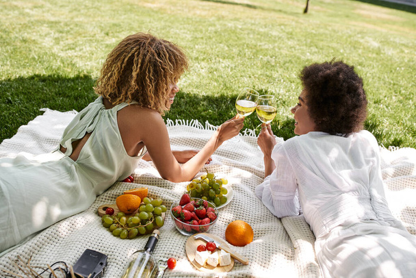 parque, picnic, novias afroamericanas tendidas en manta cerca de aperitivos y copas de vino tintineo - Foto, imagen
