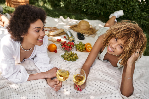 mujer afroamericana con copa de vino mirando a la cámara cerca de la novia y aperitivos en el picnic de verano - Foto, Imagen