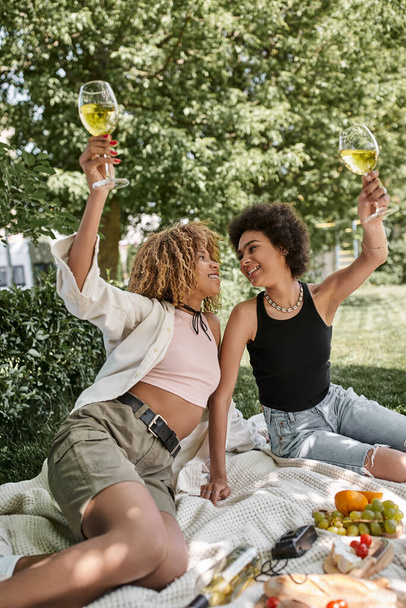 vzrušený africký Američan žena toasting s vínem sklenice poblíž přítelkyně, požitek, piknik v parku - Fotografie, Obrázek