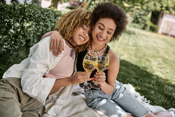positive african american girlfriends with closed eyes clinking wine glasses, summer picnic in park - Fotó, kép