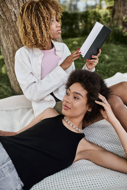 leisure in summer park, african american woman reading book near girlfriend laying on blanket - 写真・画像