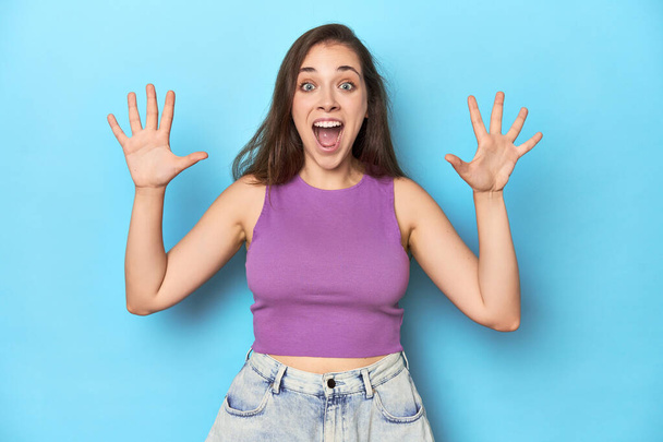 Jeune femme à la mode dans un haut violet sur fond bleu recevant une agréable surprise, excité et levant les mains. - Photo, image