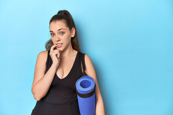 Молода спортивна кавказька жінка з йога килимок у синій студії Молода спортивна кавказька жінка з йога килимом у блакитній студії думати про щось, дивлячись на копіювальний простір. - Фото, зображення
