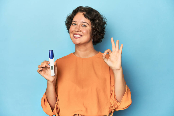 Young woman holding pregnancy test, studio background cheerful and confident showing ok gesture. - Photo, Image