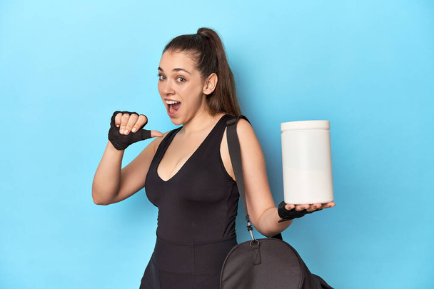 Mujer joven sosteniendo la botella de proteína en puntos de ajuste deportivo con el dedo pulgar lejos, riendo y sin preocupaciones. - Foto, imagen