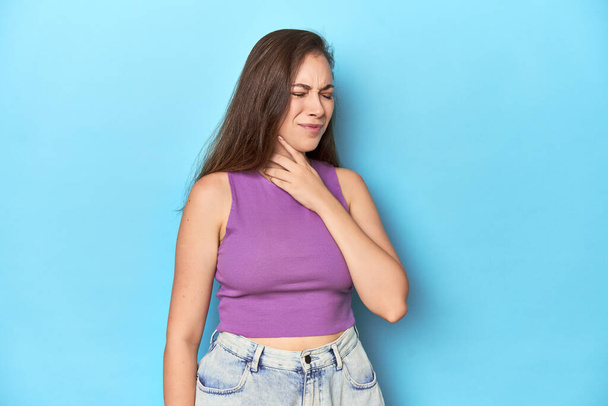 Modna młoda kobieta w fioletowej bluzce na niebieskim tle cierpi ból gardła spowodowany wirusem lub infekcją. - Zdjęcie, obraz