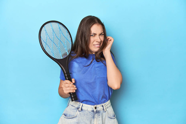 Frau mit einem elektrischen Käfer-Zapper auf blauem Hintergrund, der Ohren mit Händen bedeckt. - Foto, Bild