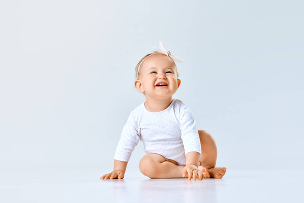 Portrait of cheerful little baby girl, toddler, child calmly sitting and laughing against grey studio background. Concept of childhood, family, newborn lifestyle, happiness, care. Copy space for ad - Zdjęcie, obraz