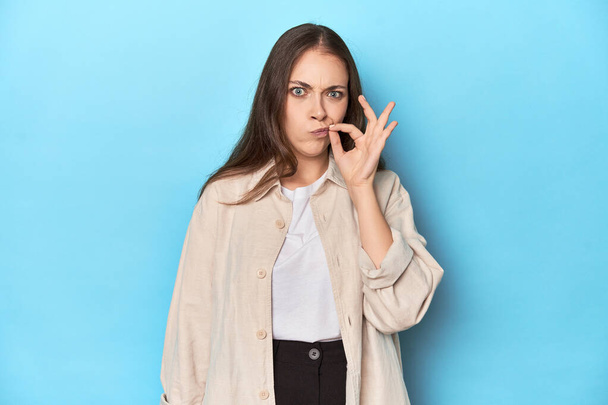 Mujer joven con estilo en una camisa sobre un fondo azul con los dedos en los labios manteniendo un secreto. - Foto, imagen
