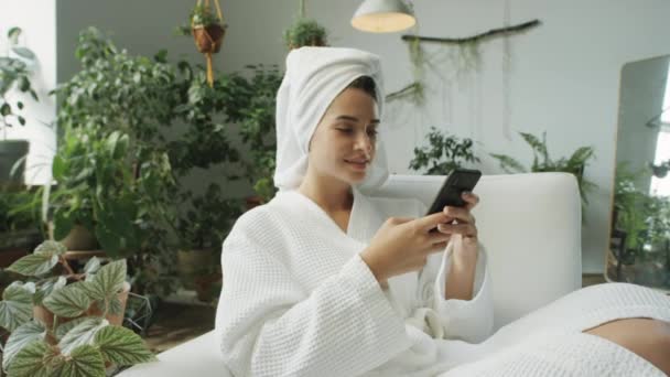Zoom σε πλάνο της όμορφης γυναίκας με μπουρνούζι κάθεται στην πολυθρόνα στο δωμάτιο διακοσμημένα με φυτά του σπιτιού και γραπτών μηνυμάτων στο κινητό τηλέφωνο - Πλάνα, βίντεο