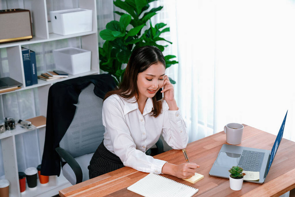 Young enthusiastic businesswoman talking on business call and writing notes on her laptop as multitask office lady. Female employee writing business tasks while talking on phone call with clients. - Photo, Image