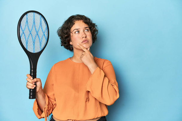電気蚊ラケットを持つ若い女性 疑わしく懐疑的な表現で横に電気蚊ラケットを見る若い女性. - 写真・画像