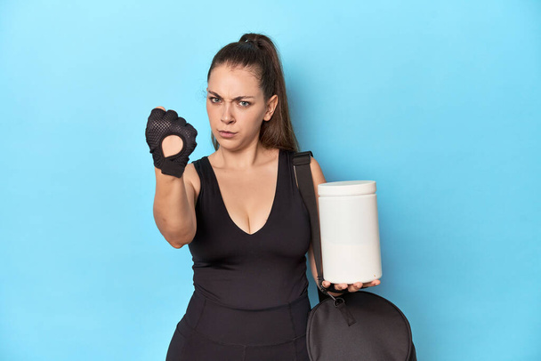 Mujer joven sosteniendo la botella de proteína en un entorno deportivo que muestra el puño a la cámara, expresión facial agresiva. - Foto, Imagen