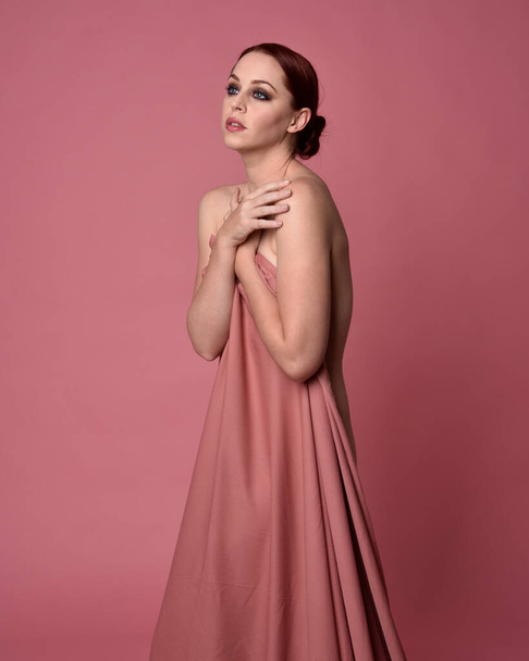  Porträt einer schönen Frau, die ein Kleid aus drapiertem Seidenstoff trägt und stehend posiert. Isoliert auf rosa Studiohintergrund. - Foto, Bild