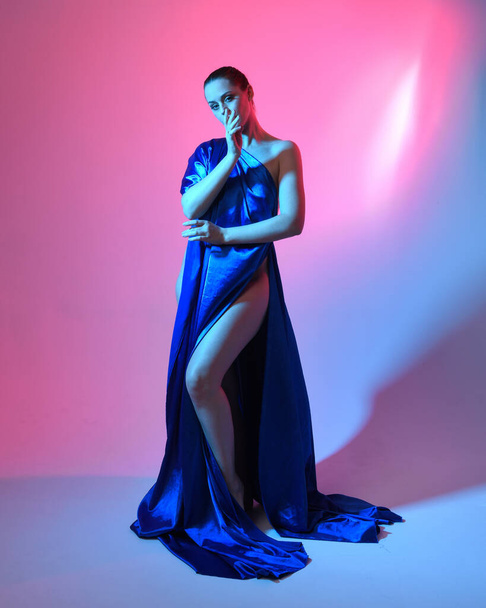 Ganztägiges Porträt der schönen weiblichen Modell trägt elegante fließende Toga Kleid aus blauer Seide drapiert, isoliert auf Studio-Hintergrund mit cineastischen bunten Beleuchtung. - Foto, Bild