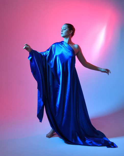 映画的なカラフルな照明でスタジオの背景に隔離された青い絹のドレープリーのエレガントな流れるガウンを身に着けている美しい女性モデルの全長の肖像画. - 写真・画像