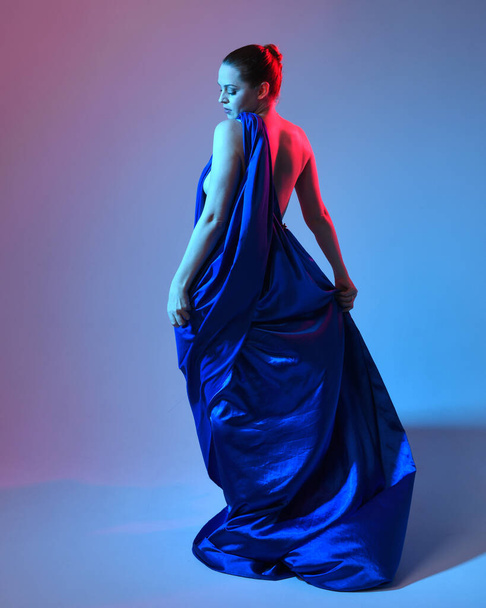 Full length πορτρέτο του όμορφου γυναικείου μοντέλου φορώντας κομψό φόρεμα που ρέει τήβεννο από μπλε μεταξωτό drapery, απομονωμένο σε φόντο στούντιο με κινηματογραφικό πολύχρωμο φωτισμό. - Φωτογραφία, εικόνα