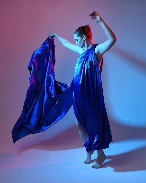 Full length portrait of beautiful female model wearing elegant flowing toga gown of blue silk drapery, isolated on studio background with cinematic colourful lighting. - Photo, Image