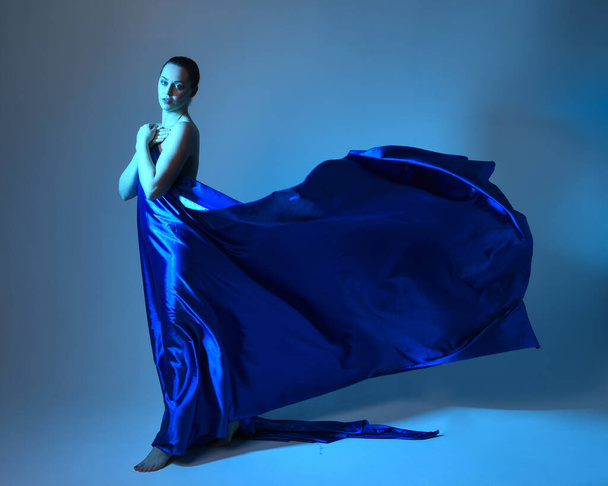Retrato de comprimento total de belo modelo feminino vestindo elegante vestido toga fluindo de cortinas de seda azul, isolado no fundo do estúdio com iluminação colorida cinematográfica. - Foto, Imagem