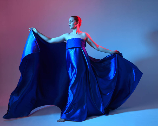 Portrait pleine longueur d'une belle mannequin féminine portant une élégante toge fluide de draperie de soie bleue, isolée sur fond de studio avec un éclairage coloré cinématographique. - Photo, image