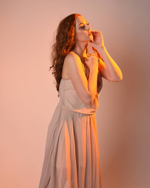 Close-up portret van mooie brunette vrouwelijke model dragen van een crème jurk met gouden achtergrondlicht verlichting.Gesturale arm poses met handen reiken, geïsoleerd op studio achtergrond. - Foto, afbeelding