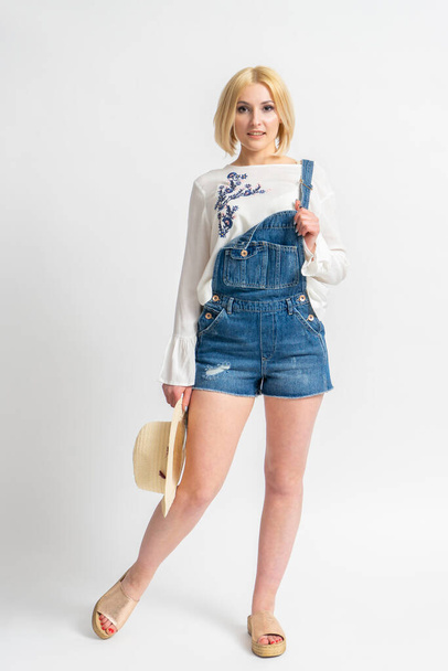Young blonde woman in casual summer denim overalls and a white rustic blouse with a hat in her hand posing on a white background - Photo, Image