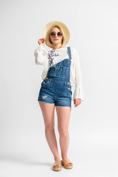 Young blonde woman wearing casual summer denim overall shorts and white rustic blouse with straw hat and sunglasses posing on white background - Фото, изображение