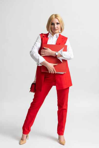 Młoda blondynka w garniturze w czerwonych spodniach z dużym folderem biurowym i damską torebką na ramieniu, pozująca na białym tle - Zdjęcie, obraz