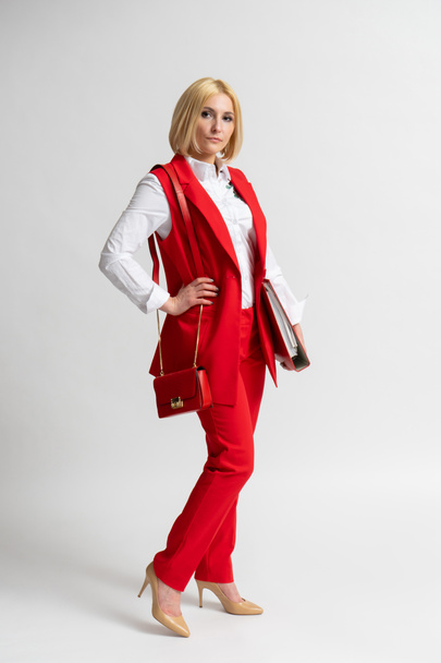 Junge blonde Geschäftsfrau in einem roten lässigen Hosenanzug mit einer großen Büromappe und einer Damenhandtasche auf der Schulter, die vor weißem Hintergrund posiert - Foto, Bild