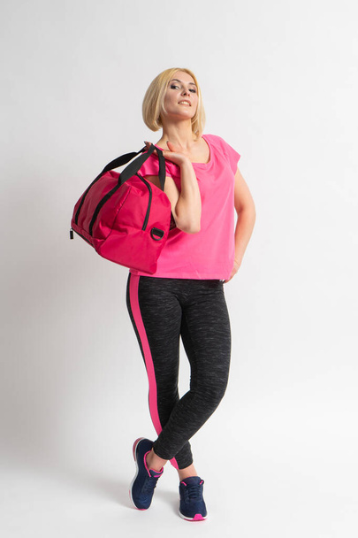 Молодая блондинка в розовой майке, спортивных леггинсах и кроссовках держит в руках большую спортивную тренировочную сумку. Фотомодель позирует в студии на белом фоне. - Фото, изображение