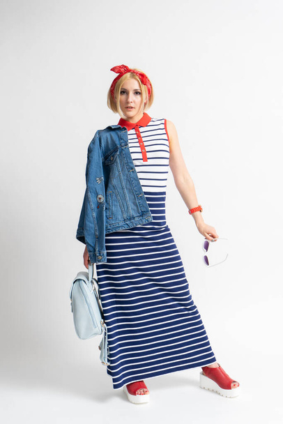 Young blonde woman in a long casual striped dress with a red bandana on her head, a backpack, sunglasses and a denim jacket in her hand posing on a white background - Photo, Image