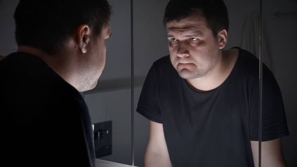 verontwaardigde en boze man in zwart t-shirt kijken in zijn reflectie op badkamer. - Foto, afbeelding