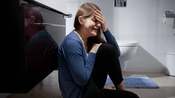 Crying woman sitting on floor in bathroom and leaning on sink. Concept of depression, home violence, suicide, stress, loneliness and frustration - 写真・画像