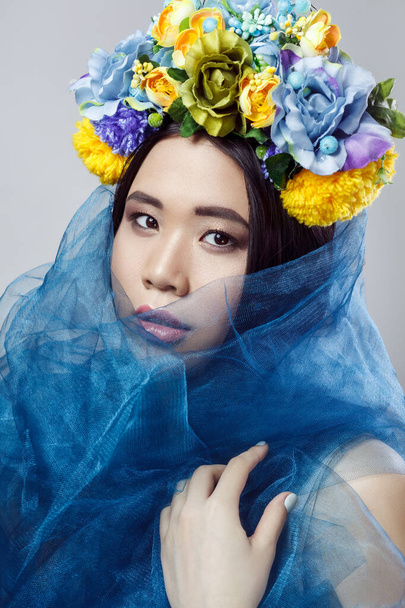 Closeup portrait of sensual beautiful woman with makeup and with floral hat and blue veil, looking at camera with serious expression. Indoor studio shot isolated on gray background - Foto, afbeelding