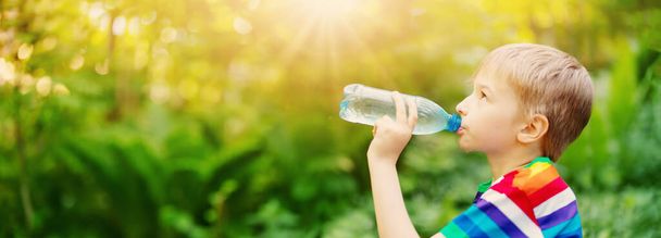 Boy standing in the natural park and drinking water from transparent bottle. Panoramic background. - Photo, image