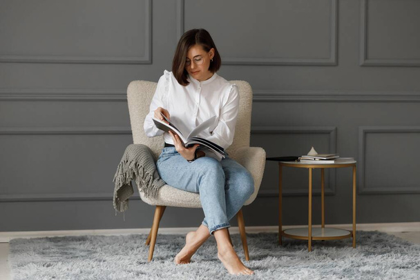 Mulher ocupada com óculos nos olhos usando uma camisa branca e jeans lendo uma revista em suas mãos sentada em uma cadeira bege com uma xadrez cinza perto de uma mesa de café em uma parede cinza no escritório - Foto, Imagem
