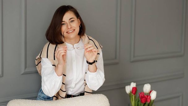Smiling businesswoman holds her glasses with both hands, wears a white shirt with a beige sweater on her shoulders, is standing leaning on a beige chair near a magazine table with a vase of tulips on it on a gray wall - Photo, Image