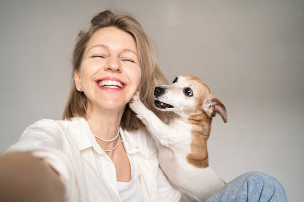 Selfie portrait of beautiful blonde smiling happy woman and her small cute dog Jack Russell terrier looking at camera and smiling. having fun wit the pet. Best friends ever! white shirt and blue jeans - Photo, image
