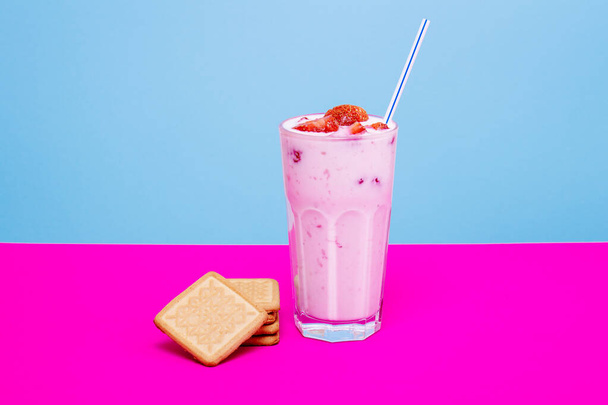 Стакан клубничного йогурта с печеньем на красочном фоне. Клубничный йогурт на голубом и розовом фоне. Стакан клубничного йогурта, покрытый свежими клубничными ломтиками.  - Фото, изображение