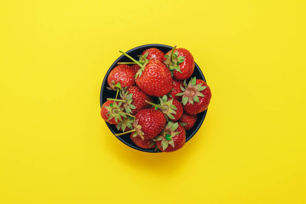 Ripe strawberry. Black bowl with strawberries on yellow background. Top view. - Zdjęcie, obraz