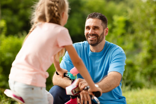 Pierwsze lekcje jazdy na rowerze. Szczęśliwy ojciec w średnim wieku uczy dziewczynkę jeździć na rowerze w parku, rozmawiać i cieszyć się spędzaniem czasu razem w wolnym czasie - Zdjęcie, obraz