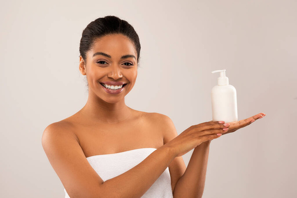 Szczęśliwy uśmiech piękna młoda czarna kobieta zawinięta biały ręcznik trzymając butelkę z produktem urody, Afryki amerykańskiej pani korzystających nawilżona skóra ciała po nowy krem, izolowane na szarym tle - Zdjęcie, obraz