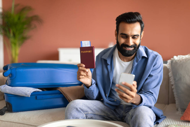 Demande de voyage. Heureux jeune homme indien utilisant un smartphone pour réserver des vacances en ligne, tenant un passeport international et des billets d'avion, assis sur le canapé près de la valise à la maison - Photo, image