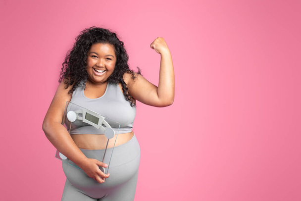 Positive black obese woman in sportswear holding scales and showing muscles biceps, standing on pink studio background, free space. Fitness, body care, strength training and weight loss - Фото, зображення