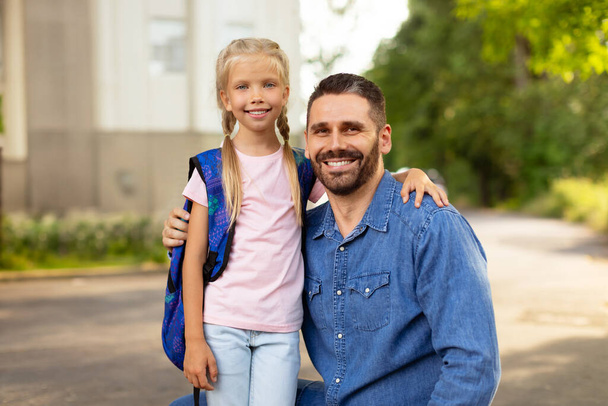 Back to school concept. Little girl with school bag embracing father near school, smiling together at camera. Happy schoolgirl with his dad on the way to school - 写真・画像